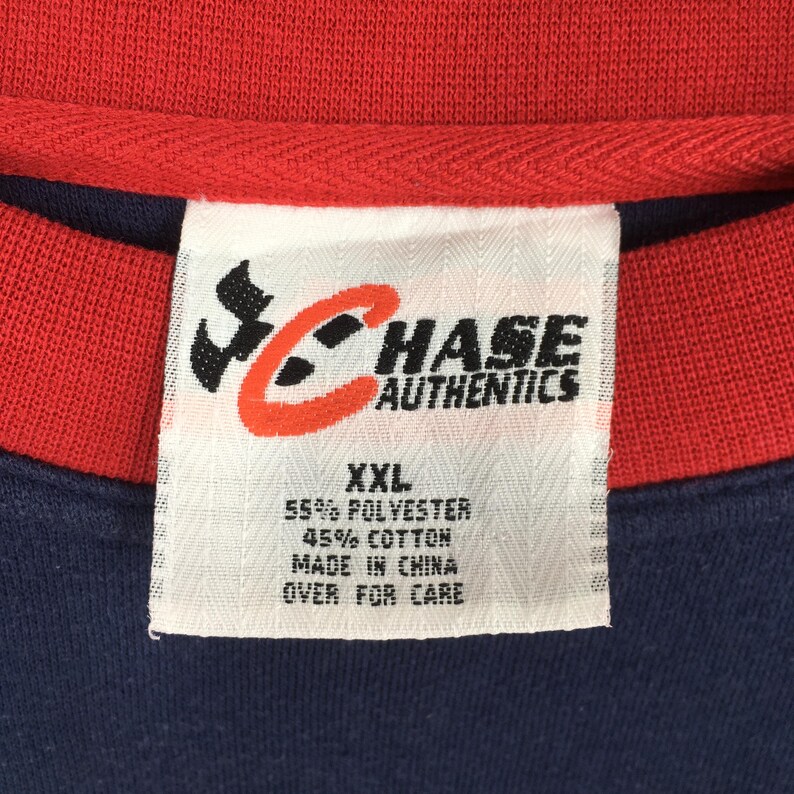 May include: Close-up of a navy blue and red shirt with a tag. The tag reads "CHASE AUTHENTICS" with a logo. The tag also indicates the shirt is XXL, made of 55% polyester and 45% cotton, and made in China.