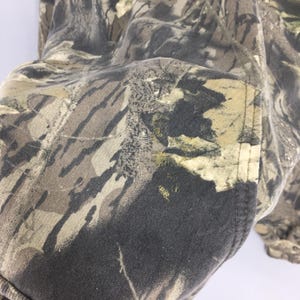 Pode incluir: Close-up de um tecido com padr&atilde;o de camuflagem. O tecido apresenta uma mistura de tons castanhos, verdes e pretos, criando um design de camuflagem florestal. A textura do tecido &eacute; vis&iacute;vel, com um detalhe de bolso na lateral.