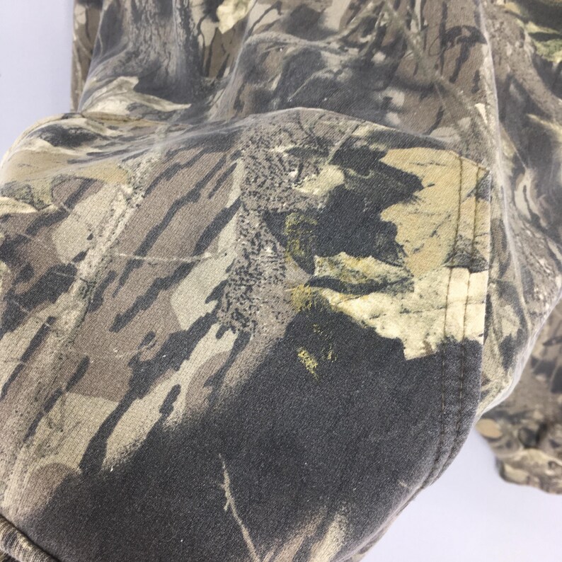 Pode incluir: Close-up de um tecido com padr&atilde;o de camuflagem. O tecido apresenta uma mistura de tons castanhos, verdes e pretos, criando um design de camuflagem florestal. A textura do tecido &eacute; vis&iacute;vel, com um detalhe de bolso na lateral.