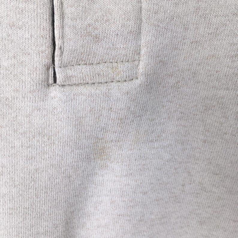 May include: Close-up of a light gray polo shirt collar. The collar features a buttoned placket and is made of a soft, textured fabric. The stitching is visible, and there are some light stains on the fabric.