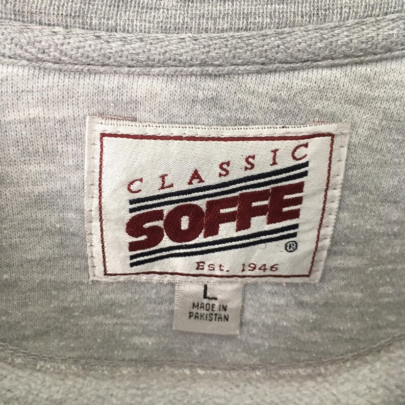 Op de afbeelding: Close-up van een grijze sweatshirt met een label met de tekst "CLASSIC SOFFE" in rode en witte letters. Het label bevat ook "Est. 1946" en "L MADE IN PAKISTAN". De sweatshirt is gemaakt van een zachte, getextureerde stof.