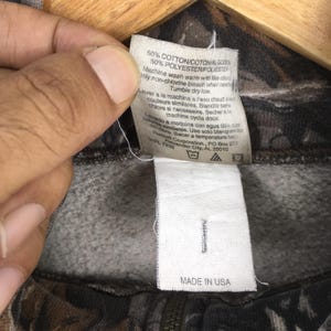May include: Close-up of a tag on a camouflage-patterned zip-up sweatshirt. The tag reads "50% COTTON/COTON/ALGODON 50% POLYESTER/POLIESTER" and includes washing instructions. The bottom of the tag reads "MADE IN USA".