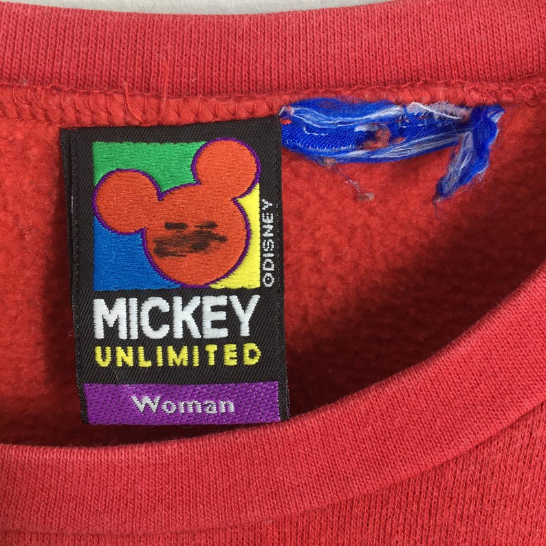 May include: Close-up of a red sweatshirt with a Mickey Unlimited label. The label features a Mickey Mouse graphic in orange, black, and yellow, with the text "MICKEY UNLIMITED" and "Woman". The label also includes the word "ODISNEY".