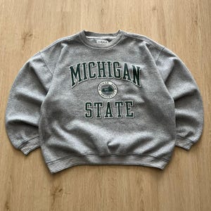 Puede incluir: Una sudadera gris de cuello redondo con las palabras "MICHIGAN STATE" en letras verdes. La sudadera tiene cuello, puños y dobladillo acanalados. La palabra "GEAR" está impresa en el cuello.