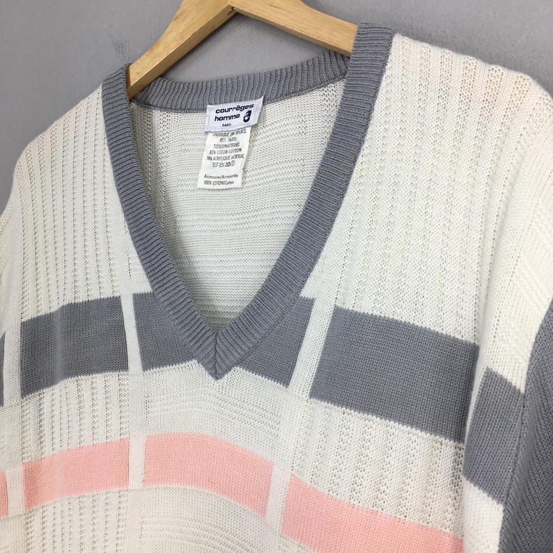 May include: A vintage ivory sweater with a V-neck and a geometric design. The sweater features gray and pink rectangular blocks. The label reads "courr&egrave;ges homme". The sweater is hanging on a wooden hanger.