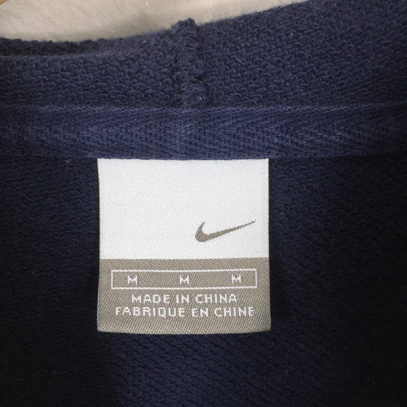 Pu&ograve; includere: Primo piano di una felpa con cappuccio blu navy con un'etichetta bianca. L'etichetta presenta il logo Nike e il testo "MADE IN CHINA" e "FABRIQUE EN CHINE". La felpa con cappuccio ha un cappuccio ed &egrave; realizzata in un tessuto strutturato.