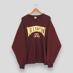 以下が含まれることがあります： 胸に黄色で「MINNESOTA」の文字がアーチ状に描かれたマルーン色のスウェットシャツ。その下に黄色の「M」ロゴがあります。スウェットシャツは木製のハンガーに吊るされています。