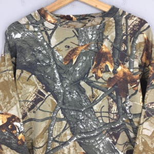 May include: A long-sleeved camouflage shirt with a crew neck. The shirt features a pattern of brown, green, and gray, with leaf and branch details. The shirt is hanging on a wooden hanger.