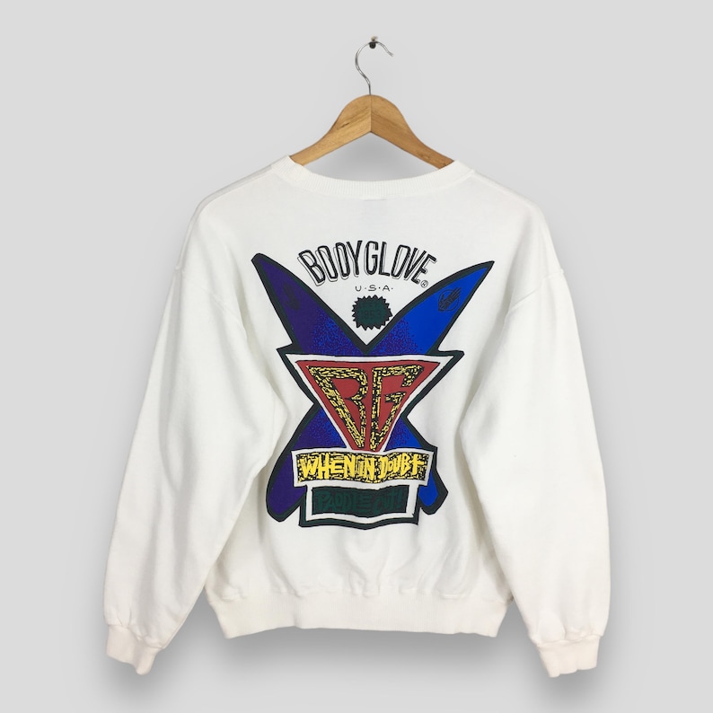 Op de afbeelding: Witte sweatshirt met een grafisch ontwerp op de rug. Het ontwerp toont de woorden "BODY GLOVE U.S.A." boven een gestileerde "X"-vorm in blauw en paars, met een rood en goud embleem in het midden. De tekst "WHEN IN DOUBT" staat eronder.