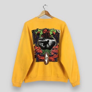 May include: Mustard yellow sweatshirt with a large graphic on the back. The graphic features a surfer silhouette, a surfboard, and text in a decorative frame. The sweatshirt has long sleeves and a crew neck.