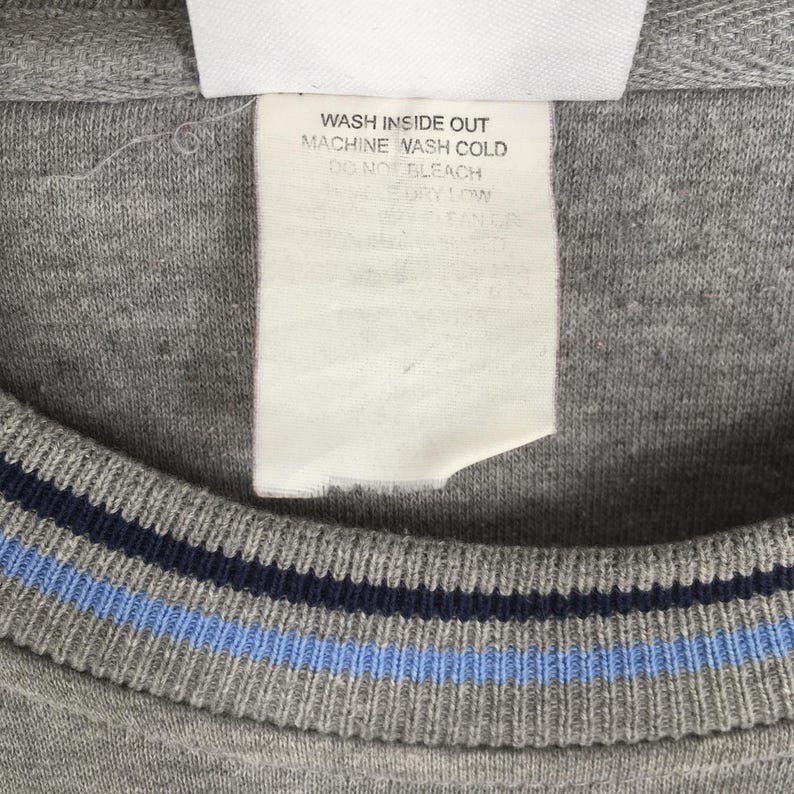 Puede incluir: Primer plano de una sudadera gris con cuello redondo y cuello a rayas azul y marino. Se ve una etiqueta blanca con texto negro, con las palabras "WASH INSIDE OUT MACHINE WASH COLD DO NOT BLEACH" impresas.