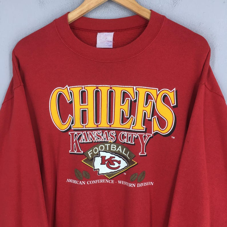 May include: Red crewneck sweatshirt featuring the word "CHIEFS" in yellow and white block letters. Below the word "CHIEFS" is "KANSAS CITY FOOTBALL" with the team logo. The sweatshirt also has the text "AMERICAN CONFERENCE - WESTERN DIVISION".