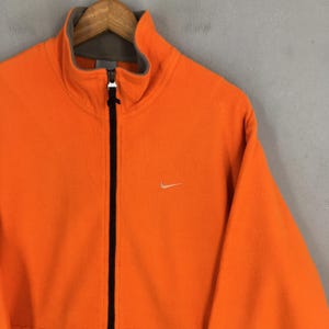 May include: Vibrant orange fleece jacket with a full-length black zip and a high collar. It has a small white Nike swoosh logo on the chest. The collar is lined with grey fabric.