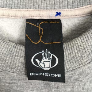 May include: Close-up of a gray sweatshirt with a black tag. The tag features a white hand graphic inside an oval and the text "BODY GLOVE" in white. The sweatshirt has a light beige collar.