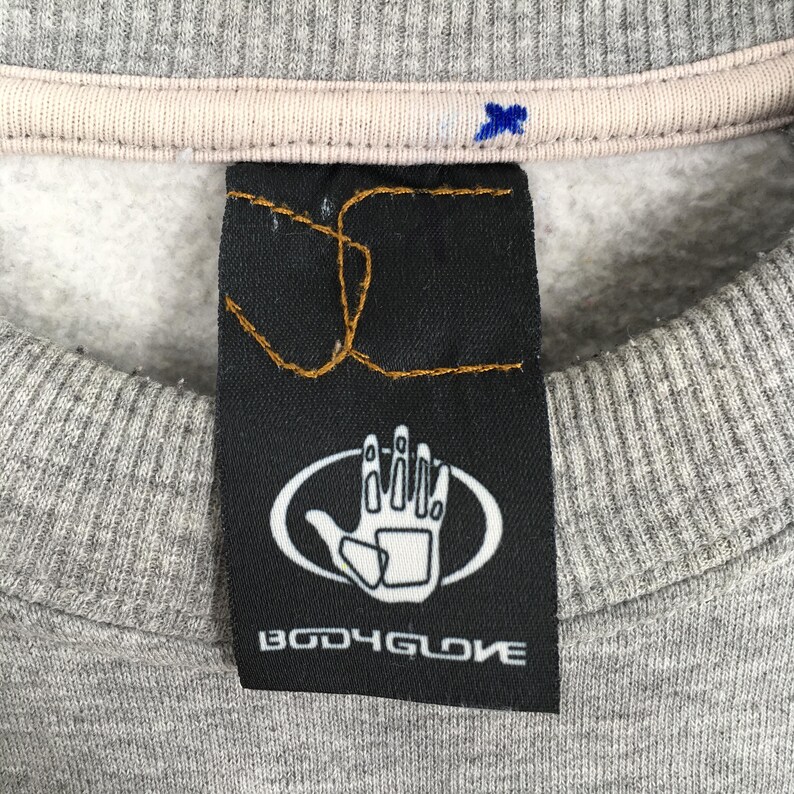 May include: Close-up of a gray sweatshirt with a black tag. The tag features a white hand graphic inside an oval and the text "BODY GLOVE" in white. The sweatshirt has a light beige collar.