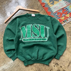 Puede incluir: Una sudadera verde con el texto "MSU SPARTANS" en letras blancas y verdes. La sudadera está colgada en una percha de madera. En el fondo se ve una alfombra colorida.