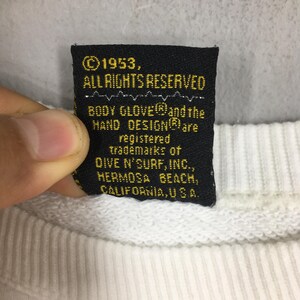 Op de afbeelding: Een zwart label met gouden tekst van een Body Glove sweatshirt. Het label luidt "&copy;1953, ALLE RECHTEN VOORBEHOUDEN" en vermeldt handelsmerken van Dive N' Surf, Inc., Hermosa Beach, Californi&euml;, VS.