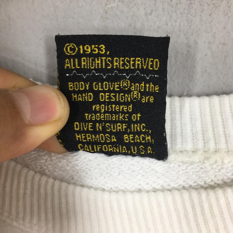Op de afbeelding: Een zwart label met gouden tekst van een Body Glove sweatshirt. Het label luidt "&copy;1953, ALLE RECHTEN VOORBEHOUDEN" en vermeldt handelsmerken van Dive N' Surf, Inc., Hermosa Beach, Californi&euml;, VS.