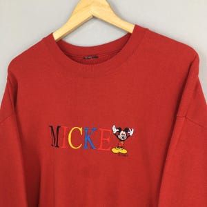 Peut inclure: Sweat-shirt rouge &agrave; col rond avec le mot "MICKEY" brod&eacute; en lettres color&eacute;es, repr&eacute;sentant le personnage de Mickey Mouse. Le sweat-shirt est suspendu &agrave; un cintre en bois.
