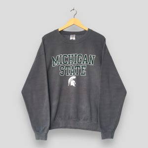 Puede incluir: Una sudadera gris carbón con las palabras "MICHIGAN STATE" en letras blancas y verdes. Debajo del texto hay un logotipo de casco espartano blanco. La sudadera está colgada de una percha de madera.