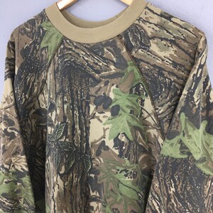Op de afbeelding: Een sweatshirt met camouflagepatroon met een beige ronde hals en raglanmouwen. Het ontwerp heeft een mix van bruine, groene en beige tinten, die een bosrijke omgeving nabootsen. Het woord "Realtree" is zichtbaar op het kledingstuk.