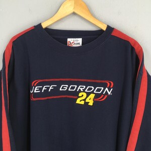 Puede incluir: Sudadera azul marino con rayas rojas en las mangas. La parte delantera muestra "JEFF GORDON 24" en rojo y amarillo. Un cuello redondo Chase Authentics vintage.