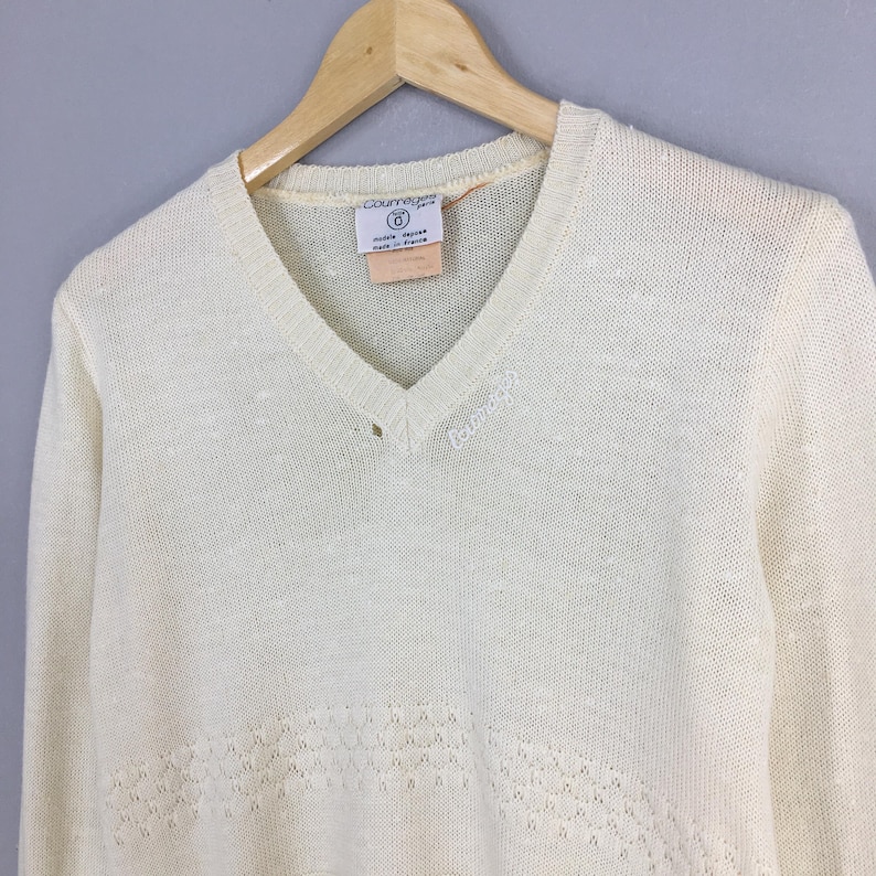 May include: Cream-colored V-neck sweater with long sleeves, hanging on a wooden hanger. The sweater features a subtle openwork design near the hem and the brand name "Courr&egrave;ges" embroidered on the chest. A tag with text is visible near the neckline.