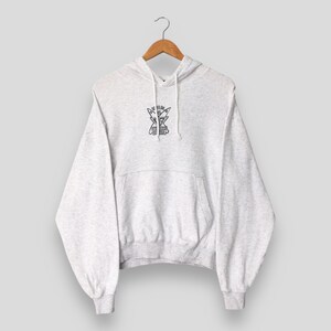 Puede incluir: Una sudadera con capucha gris claro con un bolsillo delantero y una capucha con cord&oacute;n. La sudadera tiene un peque&ntilde;o gr&aacute;fico en blanco y negro en el pecho. Cuelga de una percha de madera.