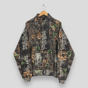 May include: A camouflage-patterned pullover sweatshirt with a quarter-zip closure. The design features a mix of brown, green, and tan colors, mimicking a forest environment. The sweatshirt has a high collar and a front pocket.