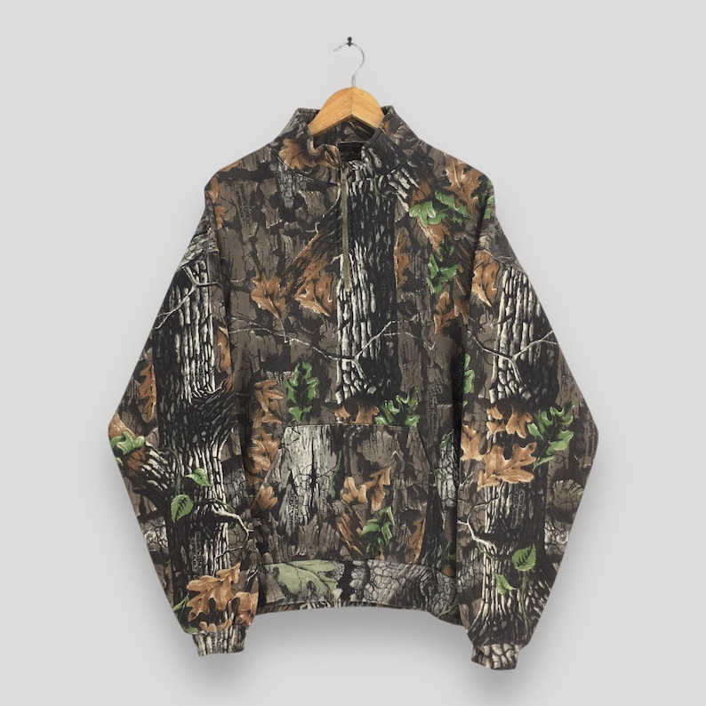 May include: A camouflage-patterned pullover sweatshirt with a quarter-zip closure. The design features a mix of brown, green, and tan colors, mimicking a forest environment. The sweatshirt has a high collar and a front pocket.