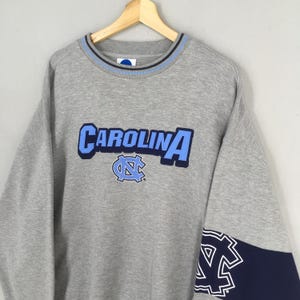 Puede incluir: Una sudadera gris con la palabra "CAROLINA" en letras azules claras y un logotipo a juego. El cuello tiene una raya azul claro y azul marino. El pu&ntilde;o de la manga es azul marino con un logotipo blanco.