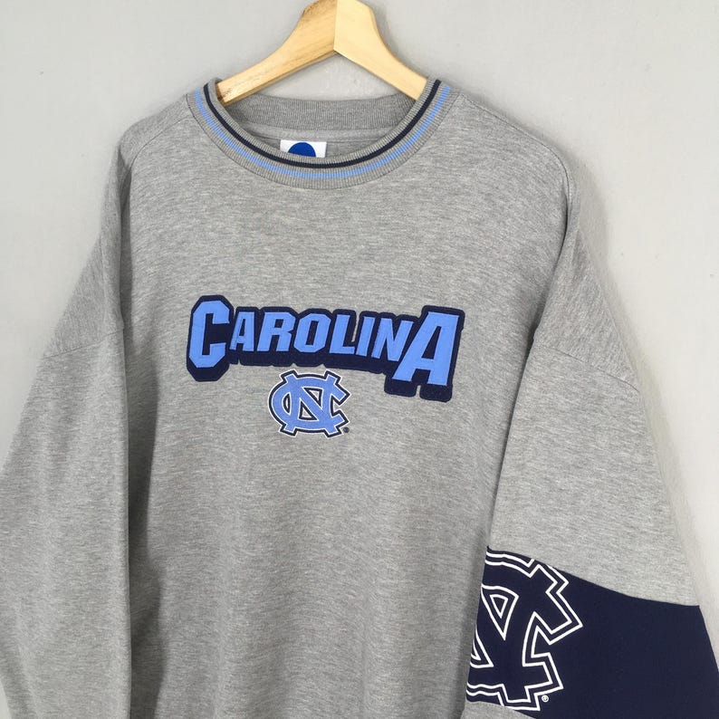 Puede incluir: Una sudadera gris con la palabra "CAROLINA" en letras azules claras y un logotipo a juego. El cuello tiene una raya azul claro y azul marino. El pu&ntilde;o de la manga es azul marino con un logotipo blanco.