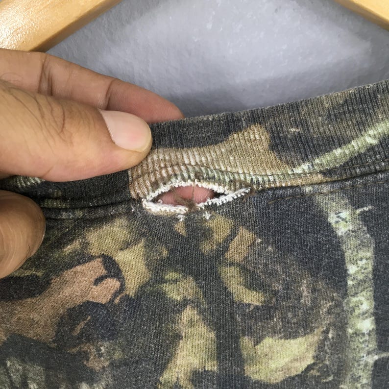 Puede incluir: Primer plano de un cuello de camisa con estampado de camuflaje con un desgarro. La tela presenta tonos de marr&oacute;n, verde y negro, t&iacute;picos de un dise&ntilde;o de camuflaje de bosque. El desgarro revela la tela interior.