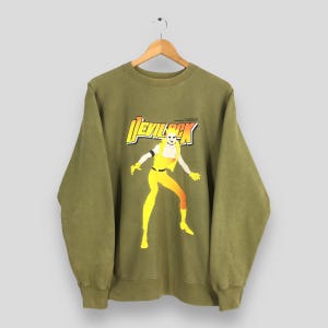 Puede incluir: Sudadera verde oliva con un gráfico de una figura amarilla con el texto "DEVIL ROCK" en amarillo y naranja. La figura lleva un atuendo amarillo con sombras naranjas. La sudadera está colgada en una percha de madera.