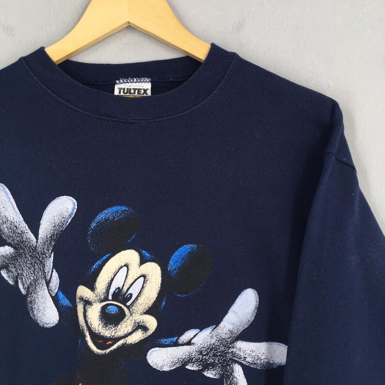 May include: Navy blue sweatshirt featuring a smiling Mickey Mouse graphic. The graphic shows Mickey with his arms outstretched. The sweatshirt has a crew neck and long sleeves. The brand tag "TULTEX" is visible at the neckline.