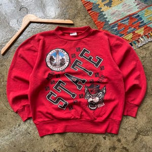 Puede incluir: Una sudadera roja con la palabra "STATE" en letras de bloque negras y blancas. La sudadera presenta un emblema circular con "NORTH CAROLINA STATE UNIVERSITY" y un gráfico de mascota de lobo. Una percha de madera está en el fondo.