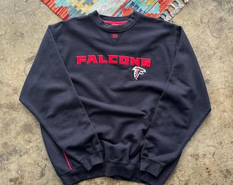 May include: A navy blue sweatshirt with the word "FALCONS" in red block letters across the chest, along with a red and white falcon logo. The sweatshirt has a red neckline and a red stripe down the side.