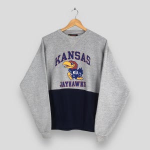 May include: A gray and navy blue sweatshirt with the word "KANSAS" arched across the chest in blue lettering. Below the word "KANSAS" is a cartoon Jayhawk mascot. The sweatshirt is hanging on a wooden hanger.