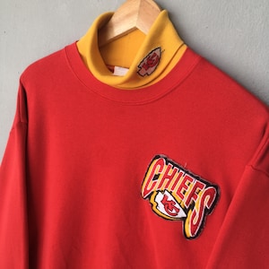 Peut inclure: Un sweat-shirt rouge dot&eacute; d'un col montant jaune avec un petit logo brod&eacute; des Kansas City Chiefs. L'avant du v&ecirc;tement pr&eacute;sente un grand &eacute;cusson cousu avec le mot CHIEFS en lettres grasses au-dessus du logo en forme de pointe de fl&egrave;che.