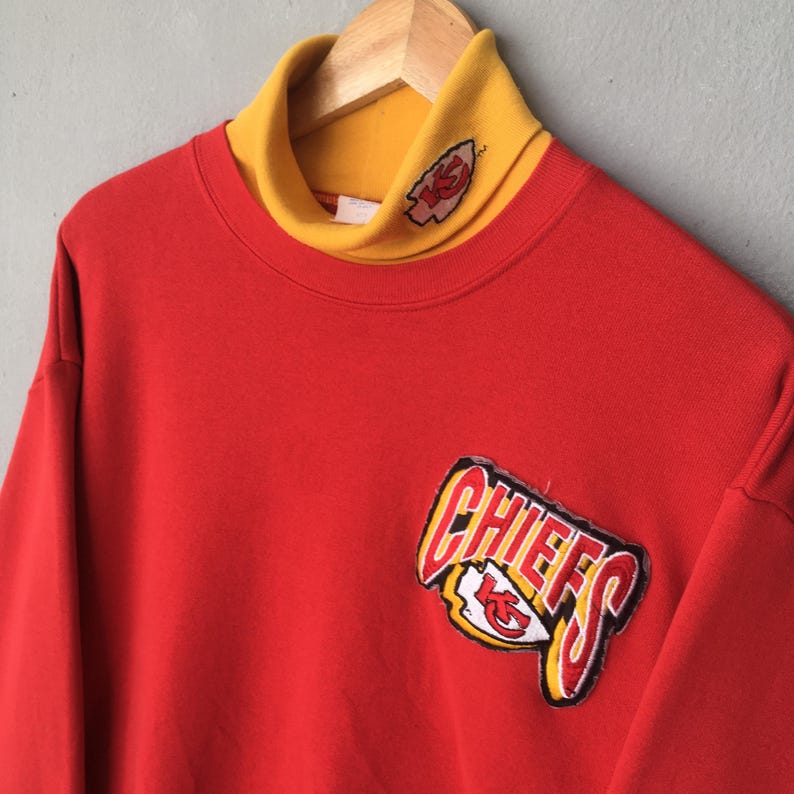 Peut inclure: Un sweat-shirt rouge dot&eacute; d'un col montant jaune avec un petit logo brod&eacute; des Kansas City Chiefs. L'avant du v&ecirc;tement pr&eacute;sente un grand &eacute;cusson cousu avec le mot CHIEFS en lettres grasses au-dessus du logo en forme de pointe de fl&egrave;che.