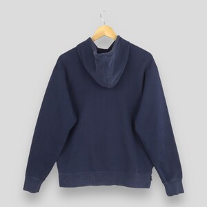 Pu&ograve; includere: Una felpa blu navy con cappuccio con coulisse, maniche lunghe e polsini e orlo a coste. La vista posteriore della felpa &egrave; esposta su una gruccia di legno su sfondo grigio chiaro. Il tessuto sembra essere un materiale morbido e usurato.