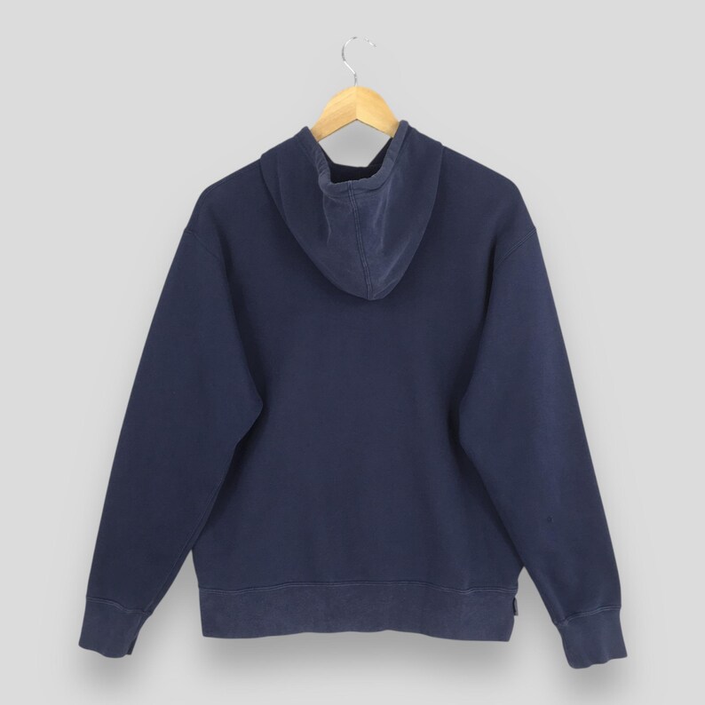 Pu&ograve; includere: Una felpa blu navy con cappuccio con coulisse, maniche lunghe e polsini e orlo a coste. La vista posteriore della felpa &egrave; esposta su una gruccia di legno su sfondo grigio chiaro. Il tessuto sembra essere un materiale morbido e usurato.