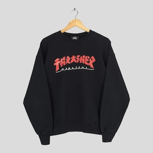 May include: Black crew neck sweatshirt featuring the word "THRASHER" in red, Japanese-style lettering across the chest. Below the word "THRASHER" is the word "magazine" in white. The sweatshirt is suspended from a wooden hanger.