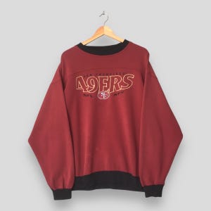 Könnte beinhalten: Bordeauxfarbenes Sweatshirt mit dem Schriftzug "SAN FRANCISCO 49ERS" in Gold. Es hat einen schwarzen Kragen, Bündchen und Saum. Die NFL- und NFC-Logos sind ebenfalls sichtbar.