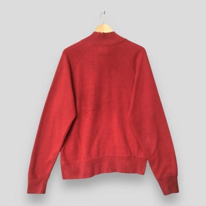 Op de afbeelding: Een effen rood sweatshirt met lange mouwen, een opstaande kraag, raglanmouwen en geribbelde boorden bij de polsen en de zoom. Het kledingstuk is gemaakt van een effen, onbedrukte stof en wordt getoond op een eenvoudige houten kledinghanger.
