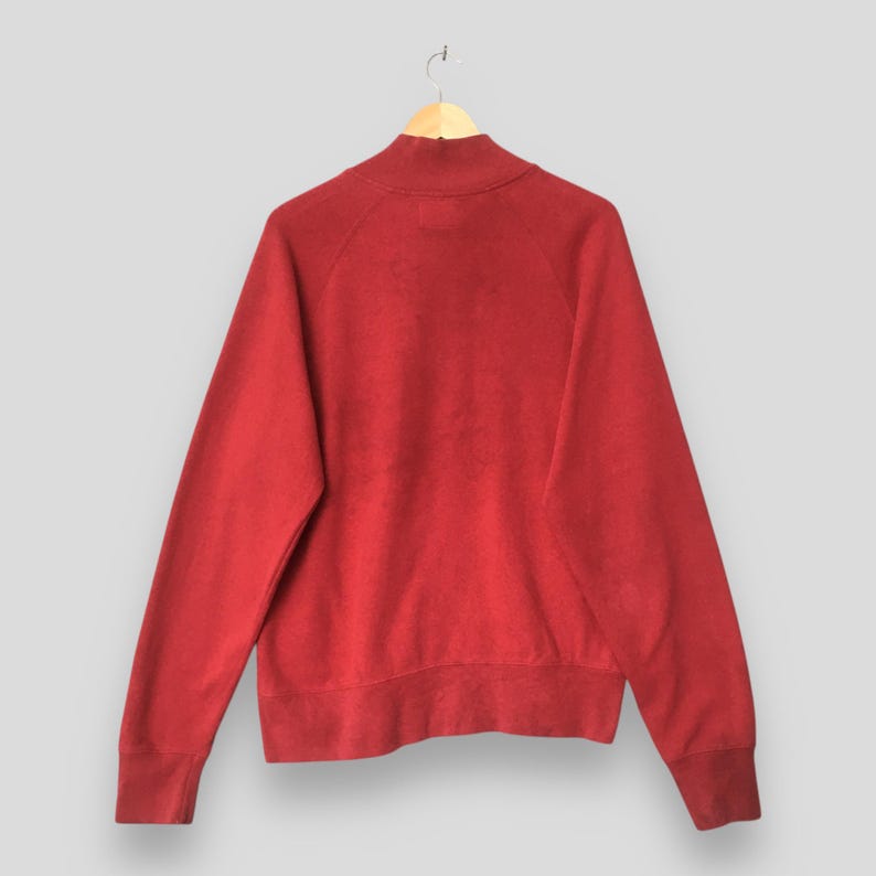 Op de afbeelding: Een effen rood sweatshirt met lange mouwen, een opstaande kraag, raglanmouwen en geribbelde boorden bij de polsen en de zoom. Het kledingstuk is gemaakt van een effen, onbedrukte stof en wordt getoond op een eenvoudige houten kledinghanger.