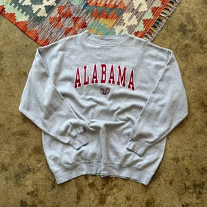 May include: A light gray sweatshirt with the word "ALABAMA" in maroon block letters across the chest. Below the word is a small maroon emblem. The sweatshirt is a crew neck style.