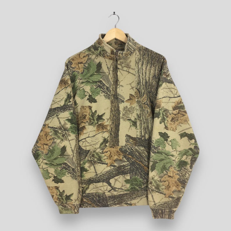 Puede incluir: Sudadera con cremallera de cuarto de vuelta con estampado de camuflaje. El dise&ntilde;o presenta una mezcla de tonos marrones, verdes y beiges, imitando un entorno forestal. La sudadera tiene un cuello alto y una cremallera frontal.