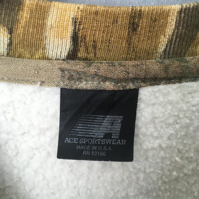 May include: Close-up of a camouflage-patterned sweatshirt collar. A black tag with the text "ACE SPORTSWEAR MADE IN U.S.A. RN 62180" is sewn into the white, fleecy interior of the garment. The collar has a brown, green, and tan pattern.