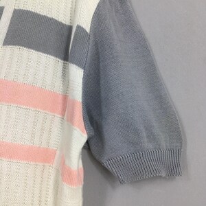 May include: Close-up of a short-sleeved knit top. The top features a color block design with white, gray, and pink horizontal stripes on the front. The sleeve is solid gray. The knit fabric has a textured appearance.